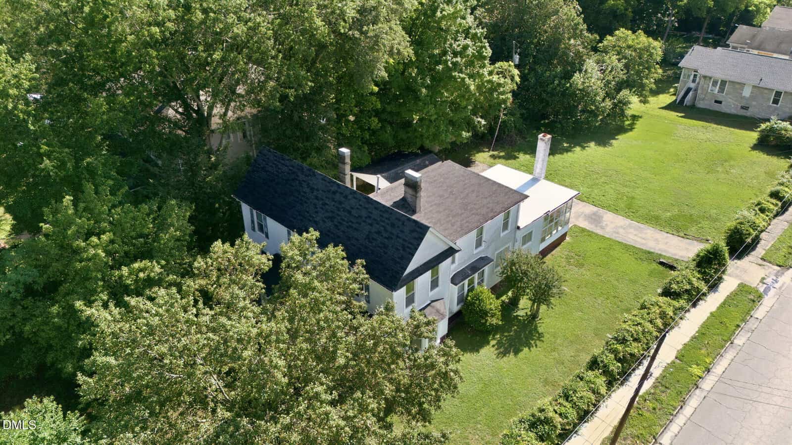 Aerial view of a two-story white house with a dark roof, surrounded by trees and green lawn, located on a corner lot with a driveway and neighboring buildings nearby.