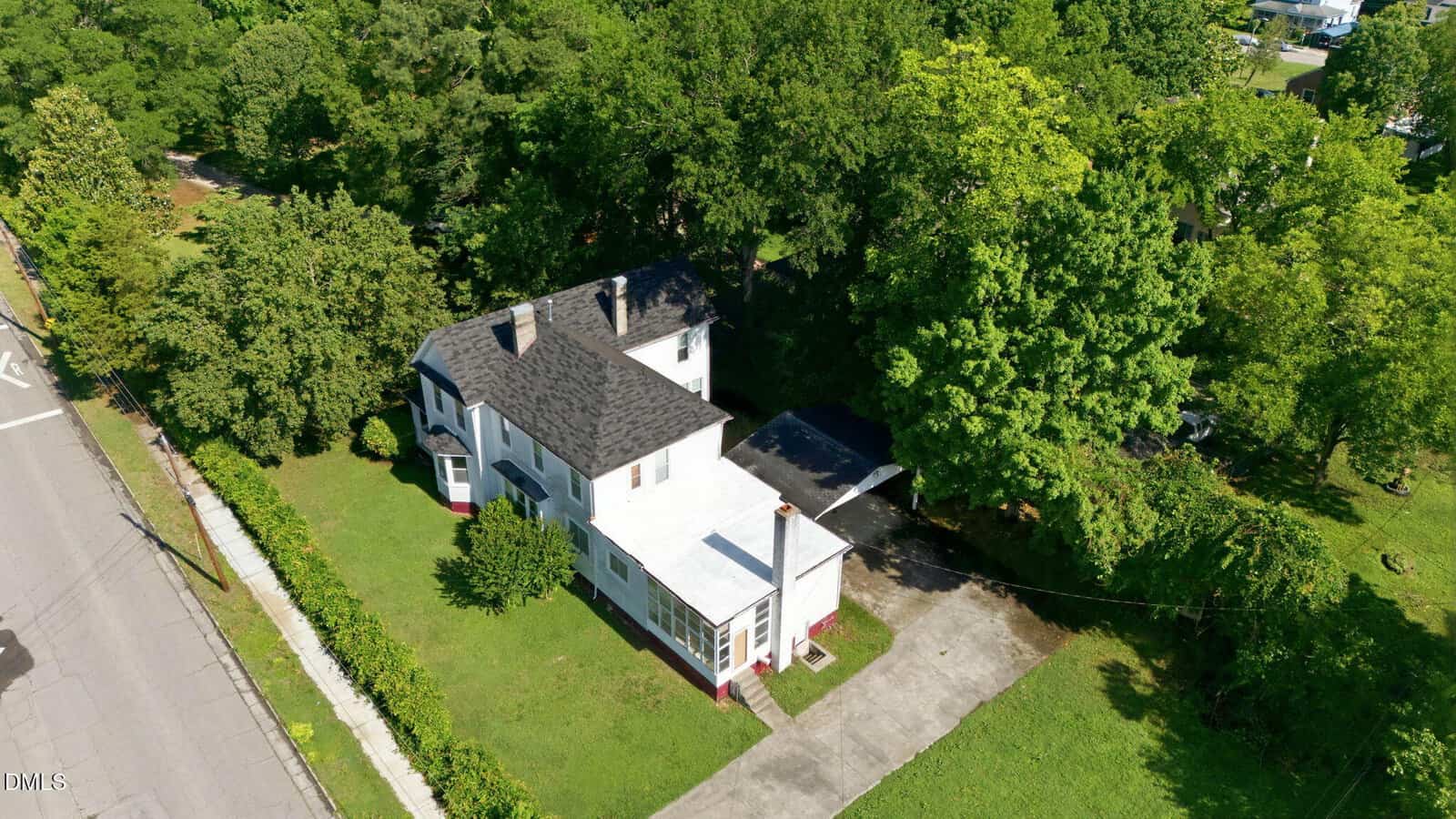 Aerial view of a two-story white house with a dark roof, driveway, carport, and a well-maintained yard surrounded by green trees along a street.