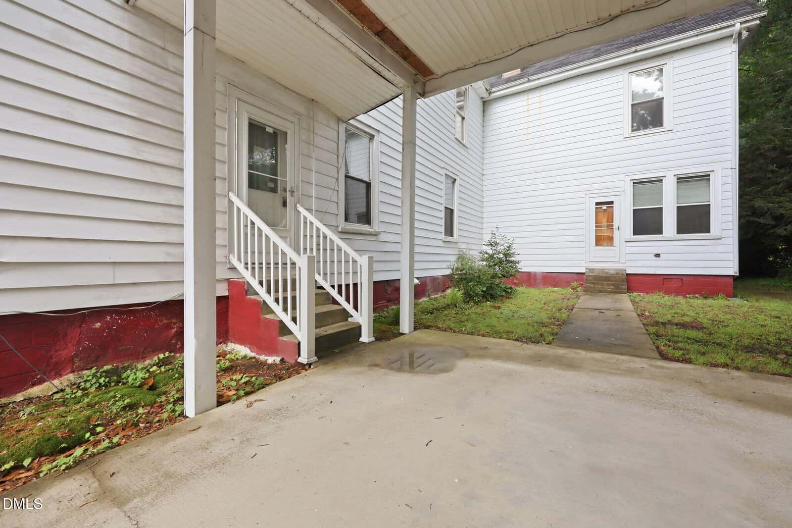 A covered concrete patio leads to a white house with red trim, featuring a small staircase with white railings and a side door. A walkway and grass area separate it from another section of the house.