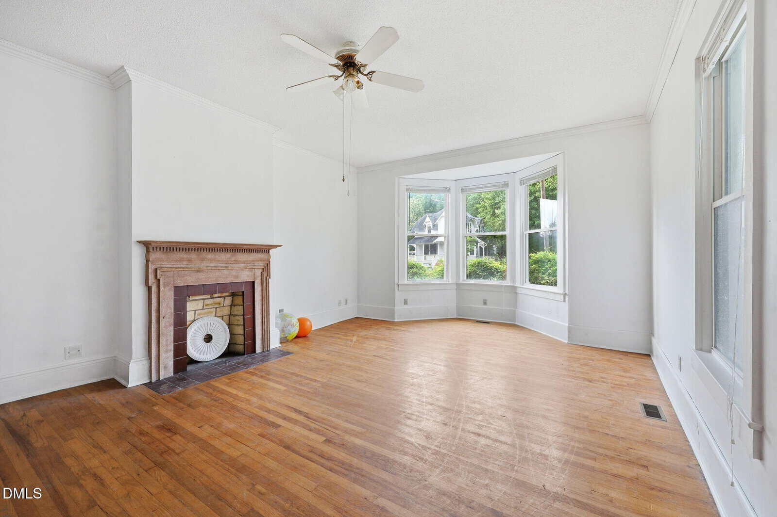 Bright, empty living room with hardwood floors, a ceiling fan, a fireplace, and large bay windows overlooking greenery; a few items sit on the hearth.