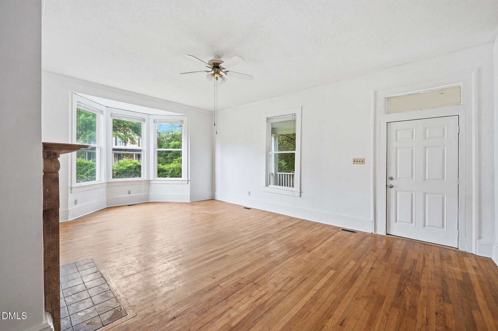 Bright, empty living room with hardwood floors, a ceiling fan, white walls, a fireplace, large bay windows, and a white door. Natural light fills the space, highlighting the open, airy feel.