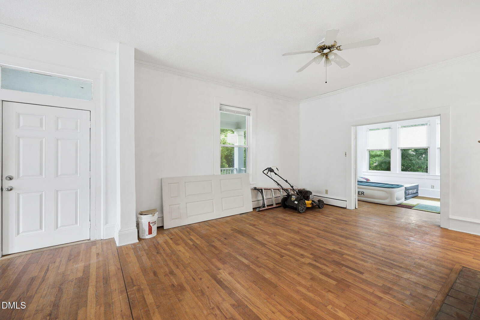 A bright empty room with hardwood floors, white walls, a ceiling fan, and a door. Against one wall are a lawnmower, a folded door, and a few tools. An adjacent room contains an inflatable mattress.