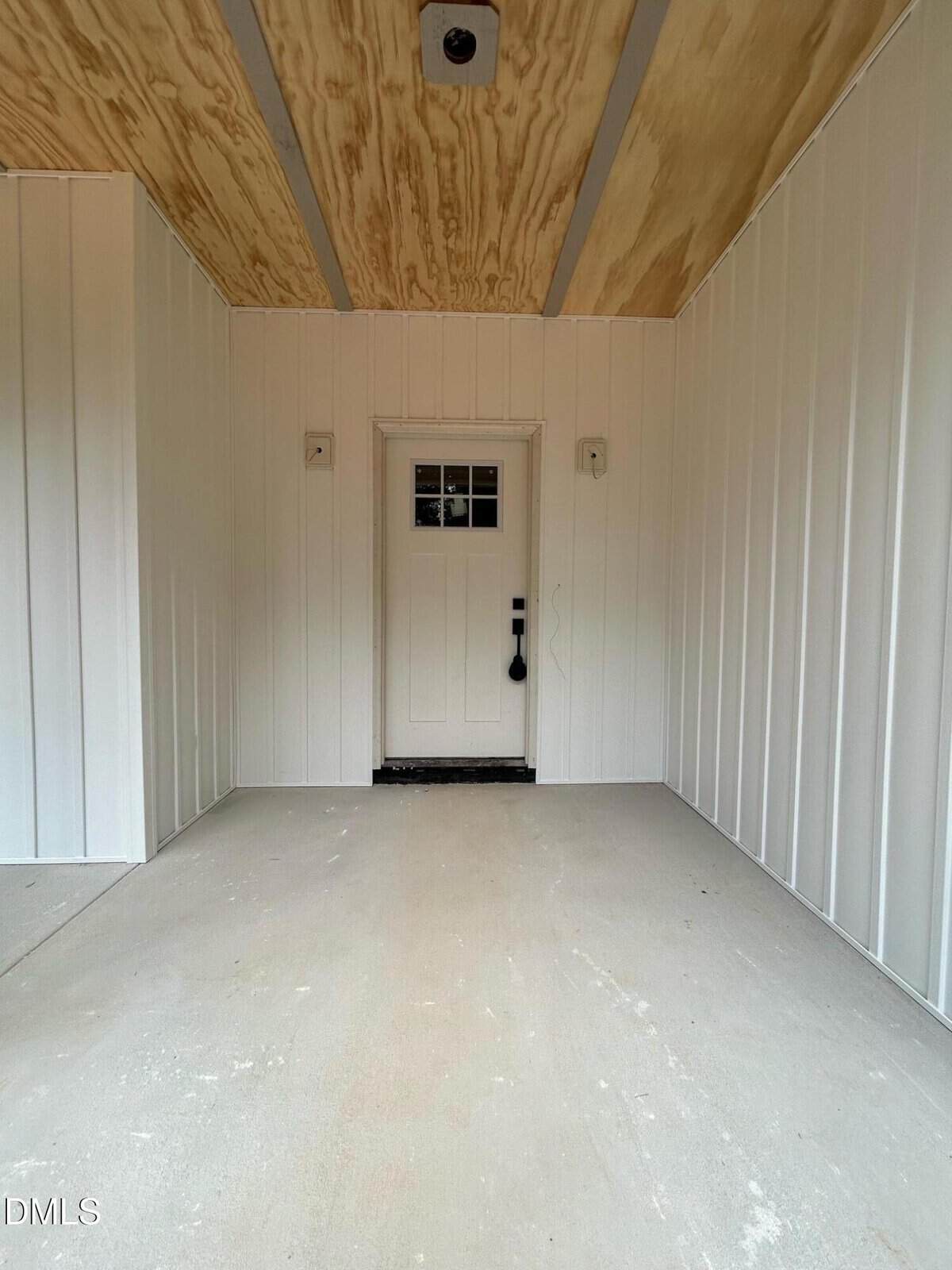 A small white door with a window panel and black handle is centered on a white paneled wall, beneath a ceiling with natural wood panels, in a clean, empty entryway or porch area.