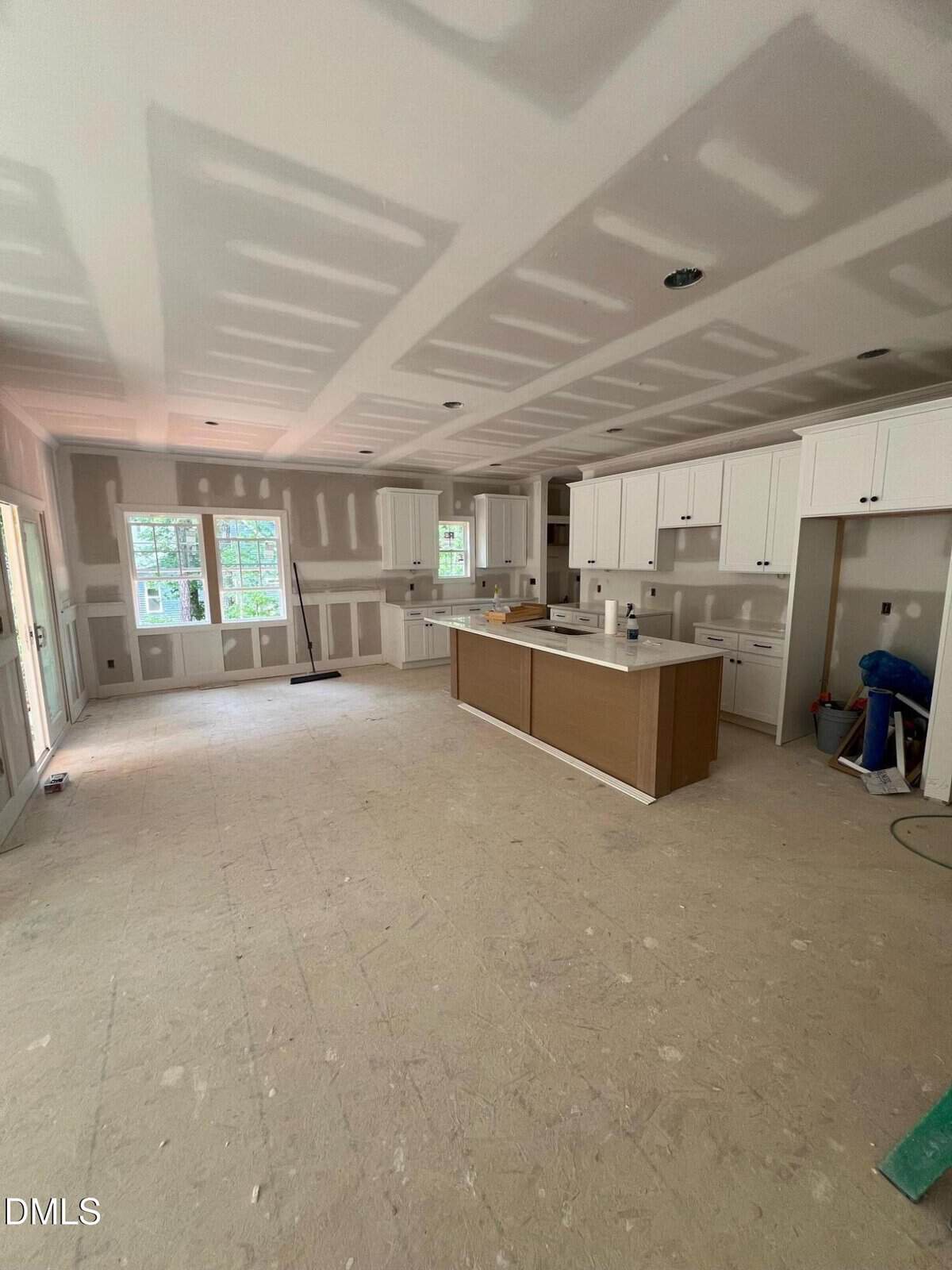 A kitchen under construction with white cabinets, a central island, unfinished drywall, and no flooring installed. Windows let in natural light, and construction materials are visible in the space.