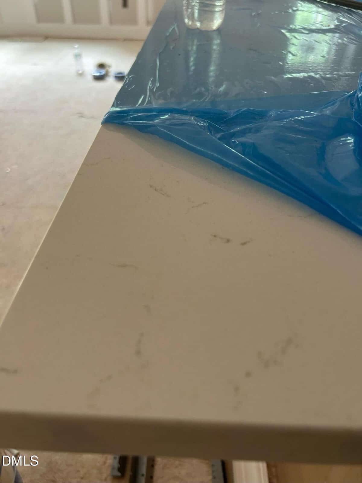A close-up of a white countertop with subtle gray veining, partially covered by blue protective plastic. The background shows a blurred room under construction with tools and materials on the floor.