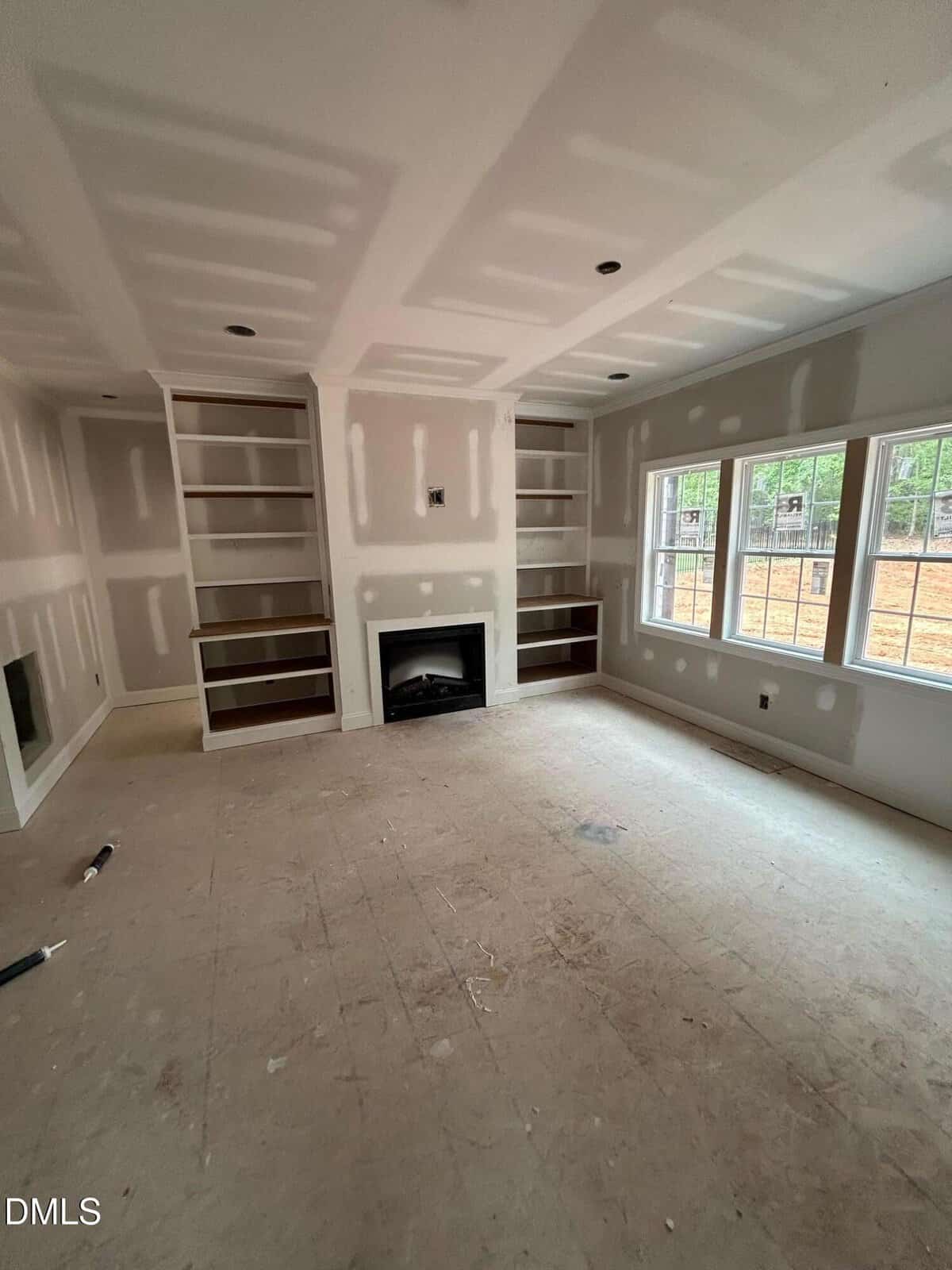 Unfinished living room with built-in shelves, a fireplace in the center, unpainted drywall, and large windows letting in natural light. Construction materials are scattered on the bare floor.