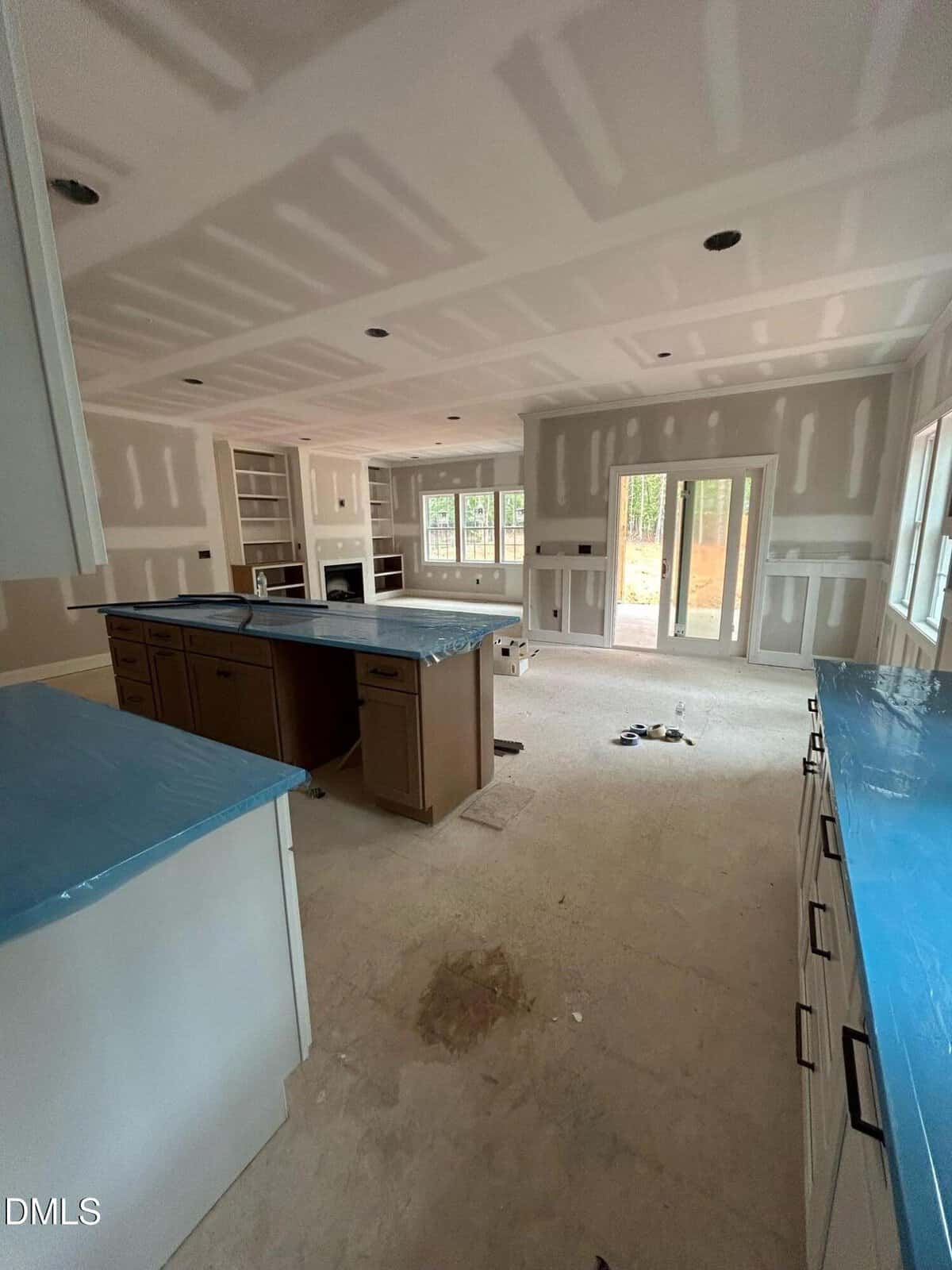 Unfinished kitchen and living area with construction materials, blue-covered countertops, unpainted walls, and large windows and doors letting in natural light.
