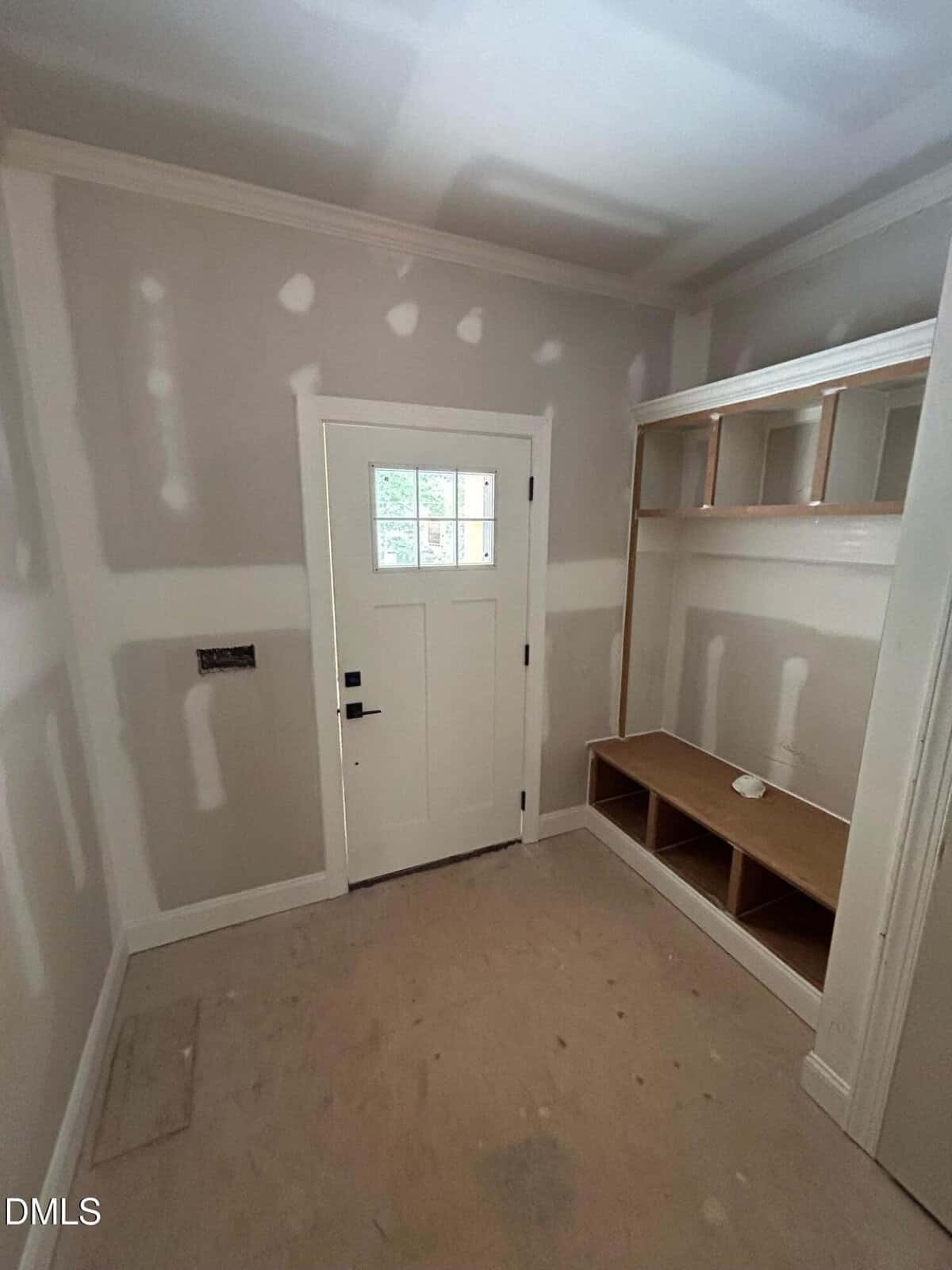 A small entryway with unfinished drywall, a white door with a window, and a built-in wooden bench with cubby holes and open shelves above. The floor is bare and construction is still in progress.