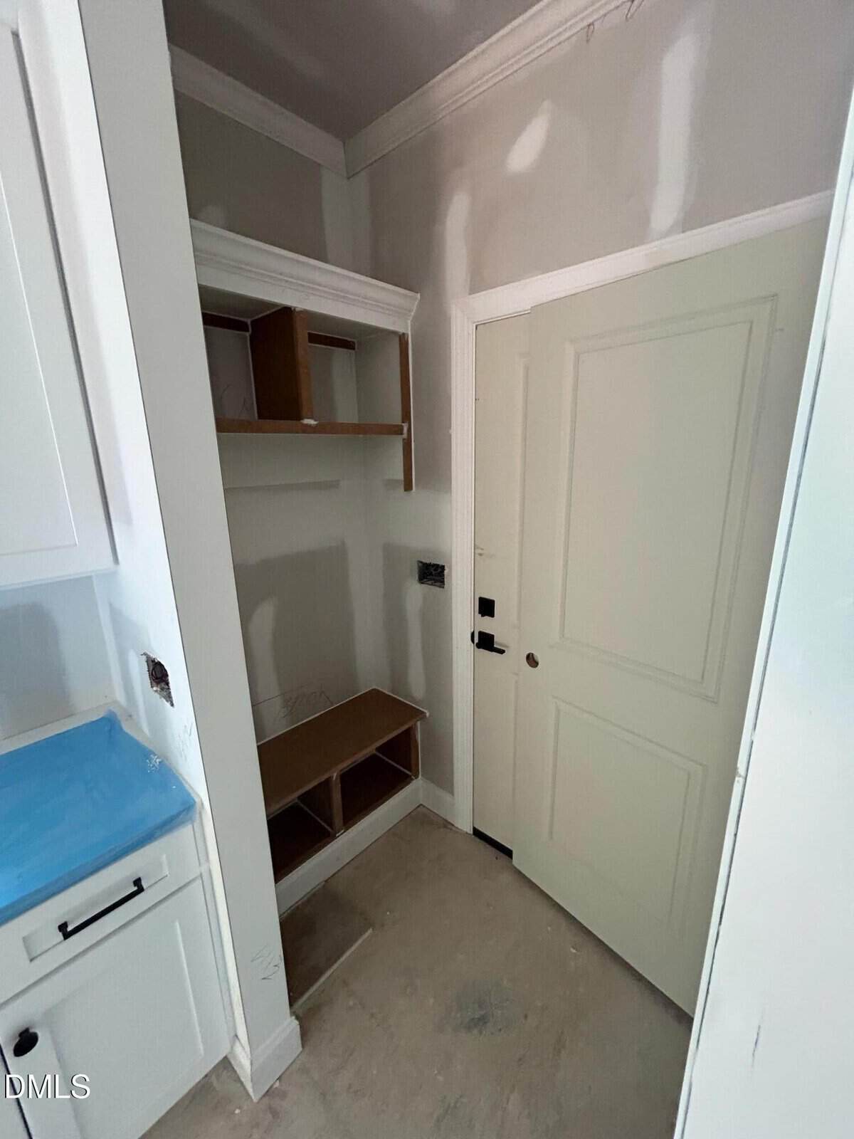 A small unfinished laundry or mudroom with unpainted drywall, a white door, built-in shelves, and a cabinet. The floor is bare, and part of the counter is covered with blue plastic.