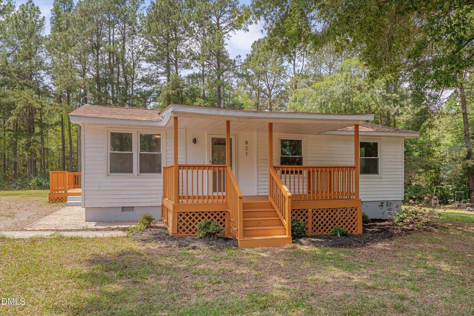 A small white house with a brown shingled roof and a wooden front porch sits at 821 Fred Royster Road, Henderson, surrounded by grass, trees, and a wooded background under a clear sky.