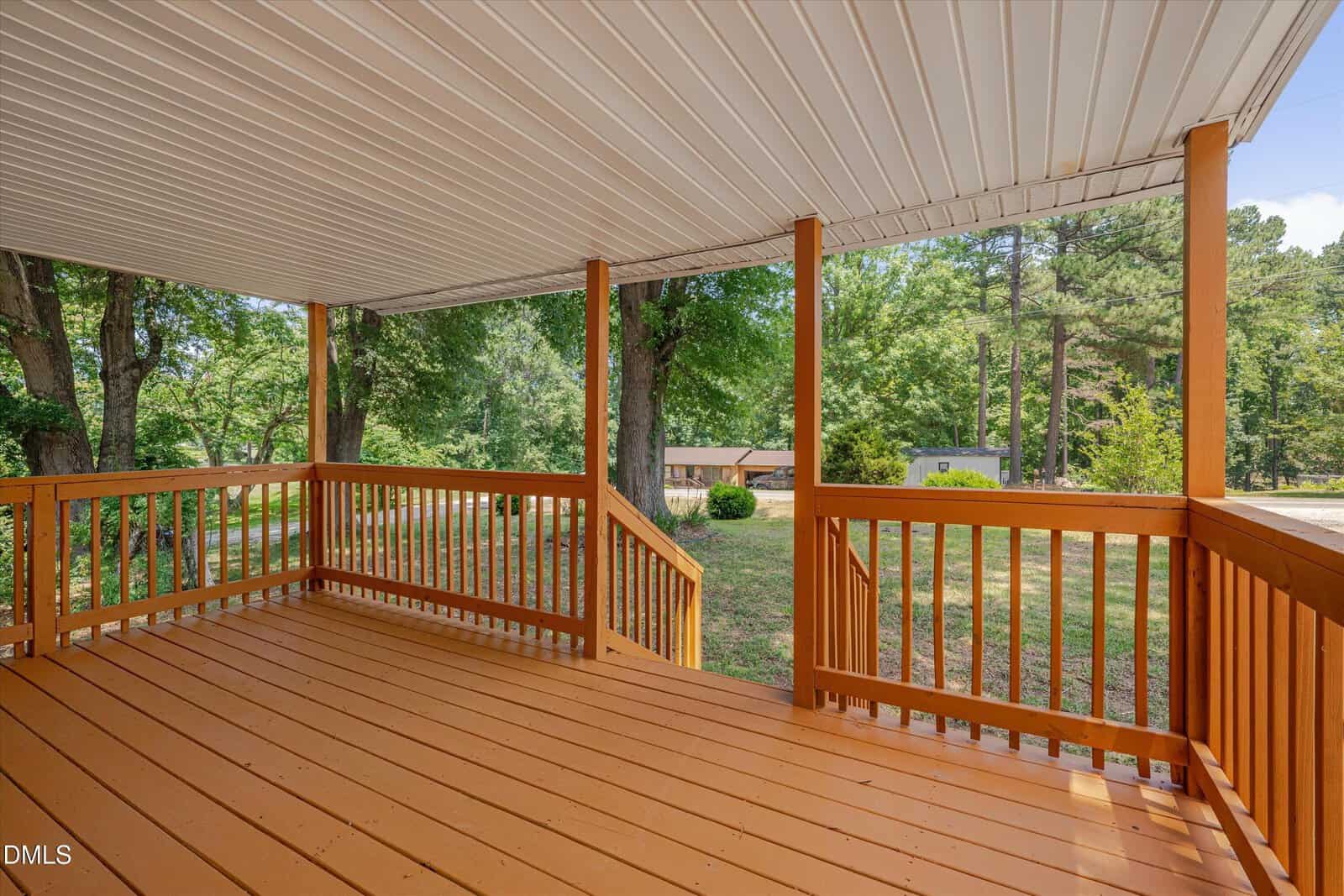 A covered wooden porch with railings overlooks a grassy yard with trees and a road in the background on a sunny day.