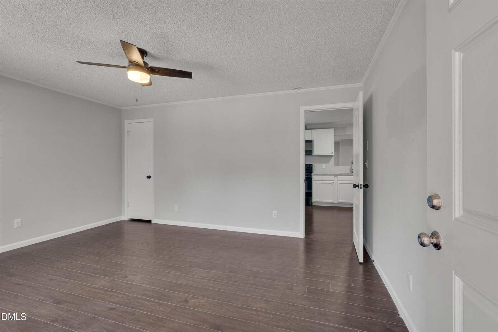 Empty room with dark wood floors, light gray walls, ceiling fan, and two doors. The room opens to a kitchen area with white cabinets and dark appliances visible in the background.