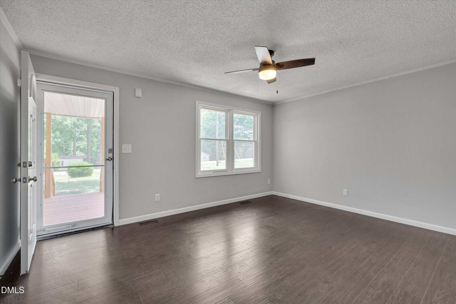 A bright, empty living room with light gray walls, a ceiling fan, two large windows, dark wood flooring, and a glass door leading to a covered porch with a view of green trees outside.