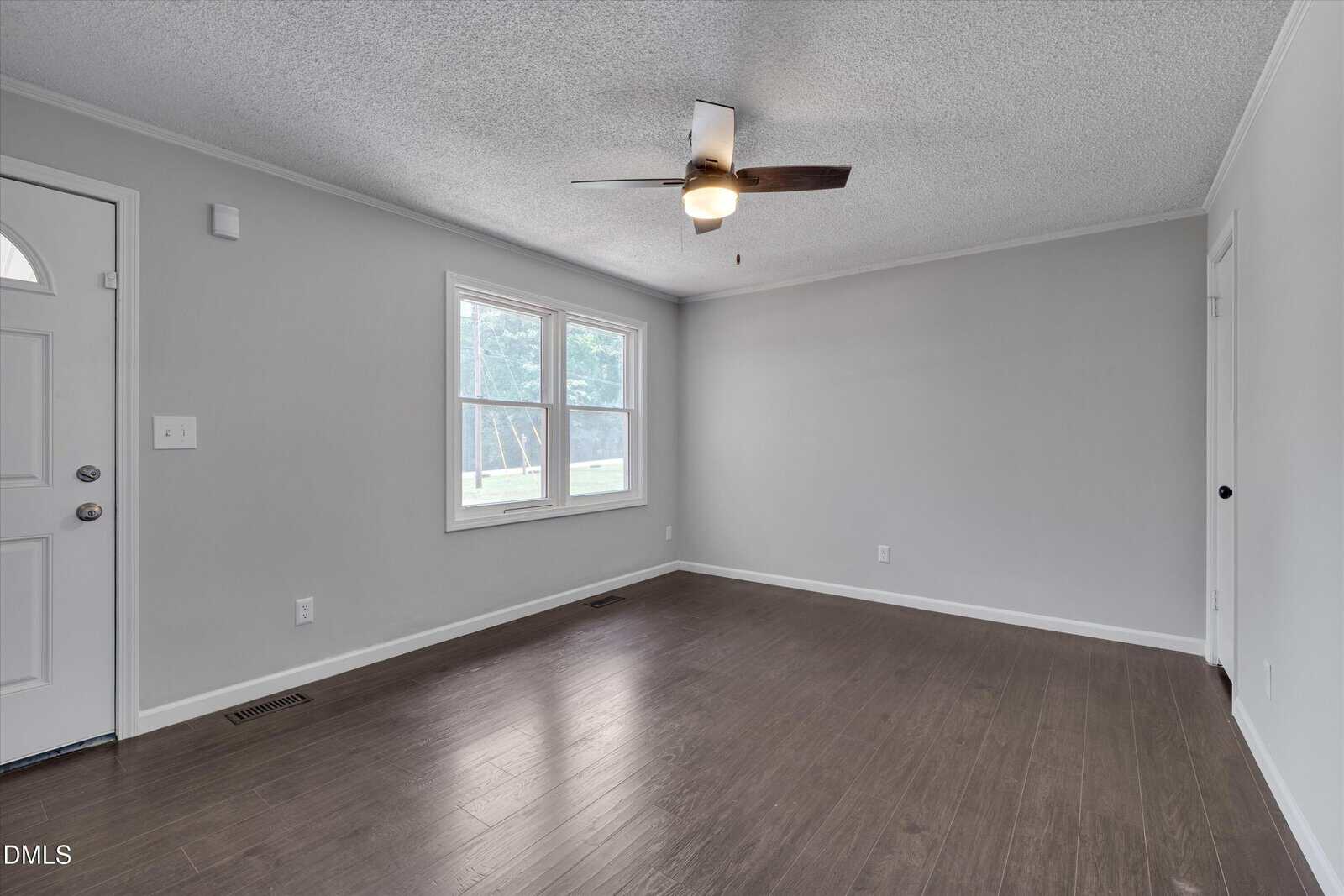Empty living room with light gray walls, dark wood flooring, a ceiling fan with a light, two large windows letting in natural light, and a white door to the left.