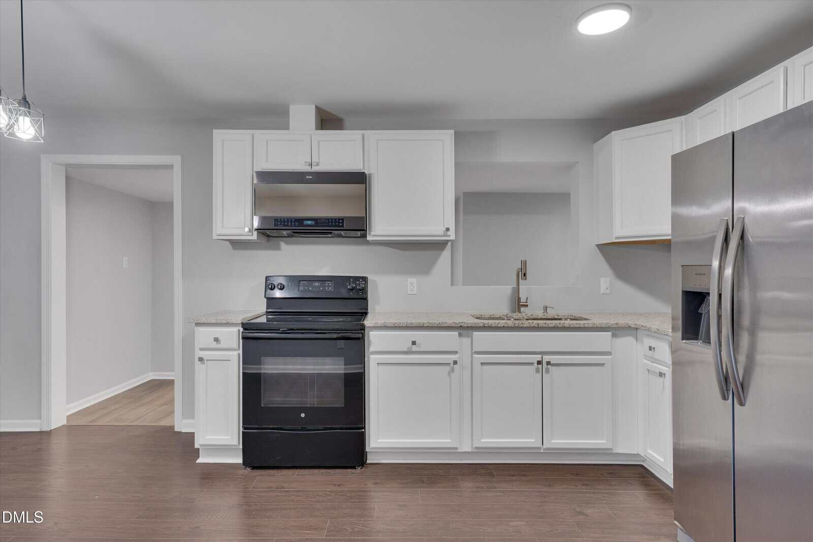 Modern kitchen with white cabinets, stainless steel appliances including a fridge, oven, and microwave, granite countertops, a sink, and dark wood flooring. Open doorway visible to the left.