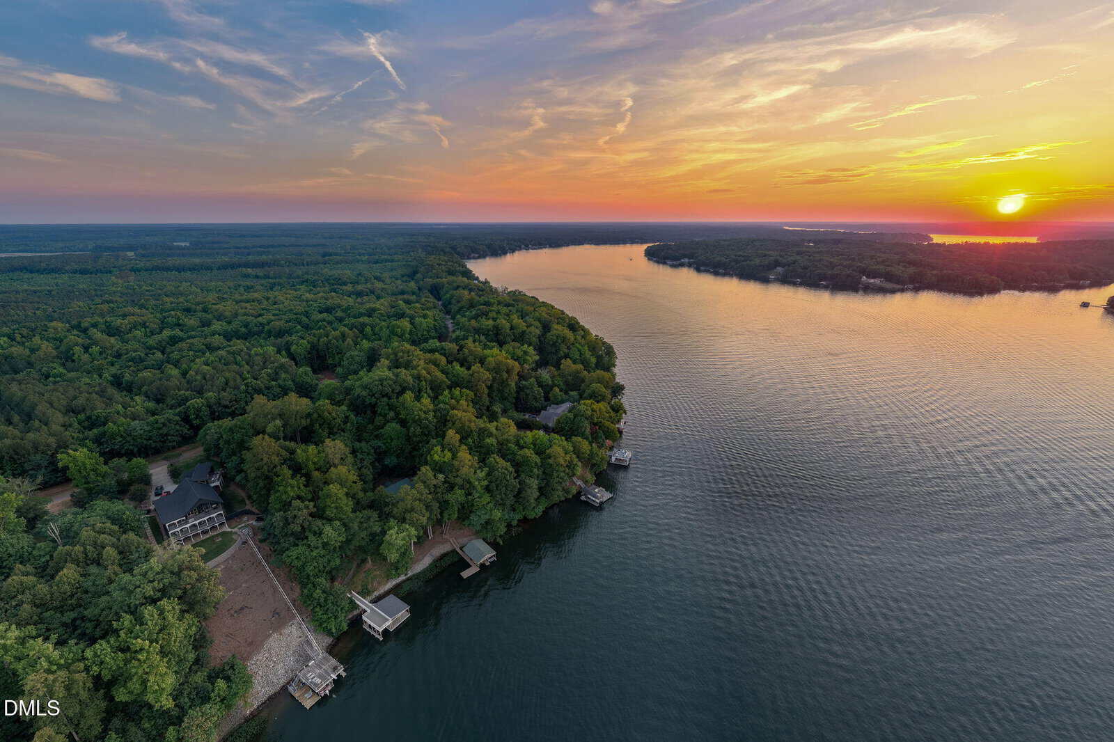 Aerial view of a sunset over a lake bordered by dense green forest, with several docks and houses along the shoreline— including Lot 15 Harbor Drive, Macon. The sky displays orange, yellow, and blue hues as the sun nears the horizon.