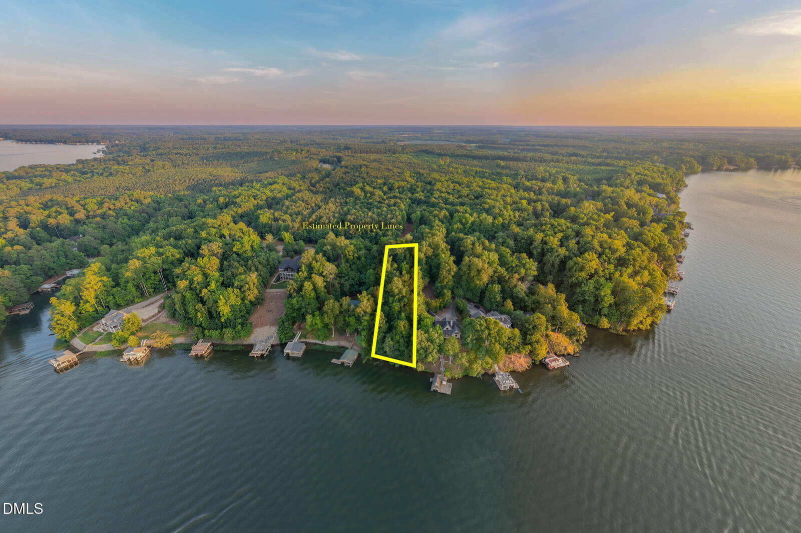 Aerial view of a wooded lakeshore with a rectangular property outlined in yellow, highlighting lakefront land surrounded by trees, docks, and other waterfront homes at sunset.
