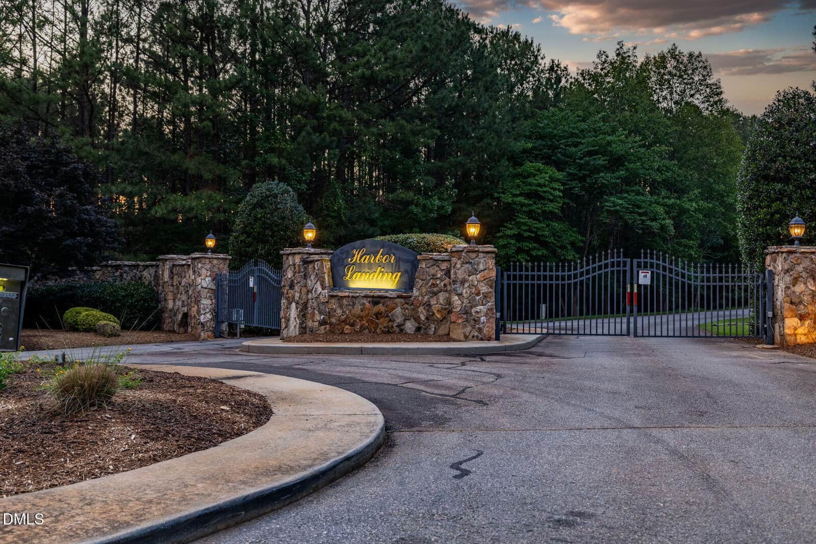 Gated entrance to Harbor Landing, with stone walls, illuminated sign, iron gate, lanterns, and trees in the background at dusk.