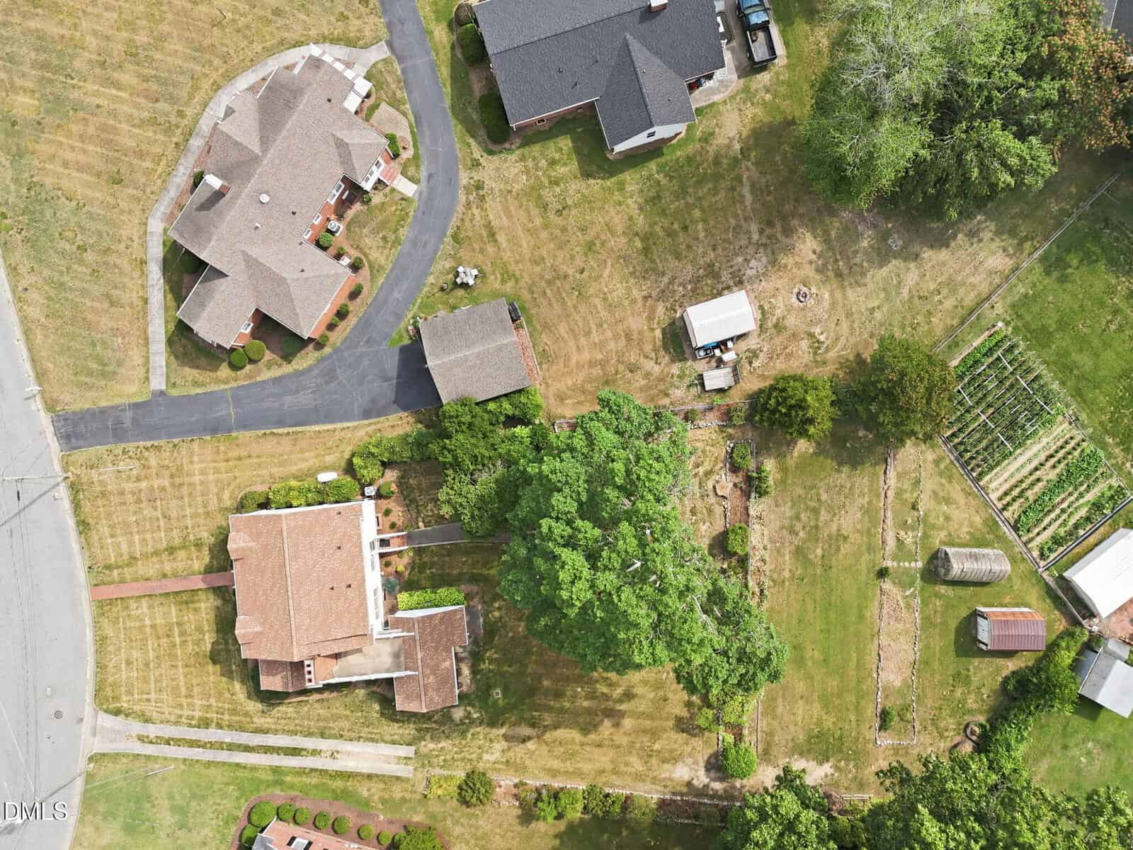 Aerial view of a residential area with two houses, driveways, outbuildings, a large tree, a garden plot, and grassy lawns separated by fences, taken on a sunny day.