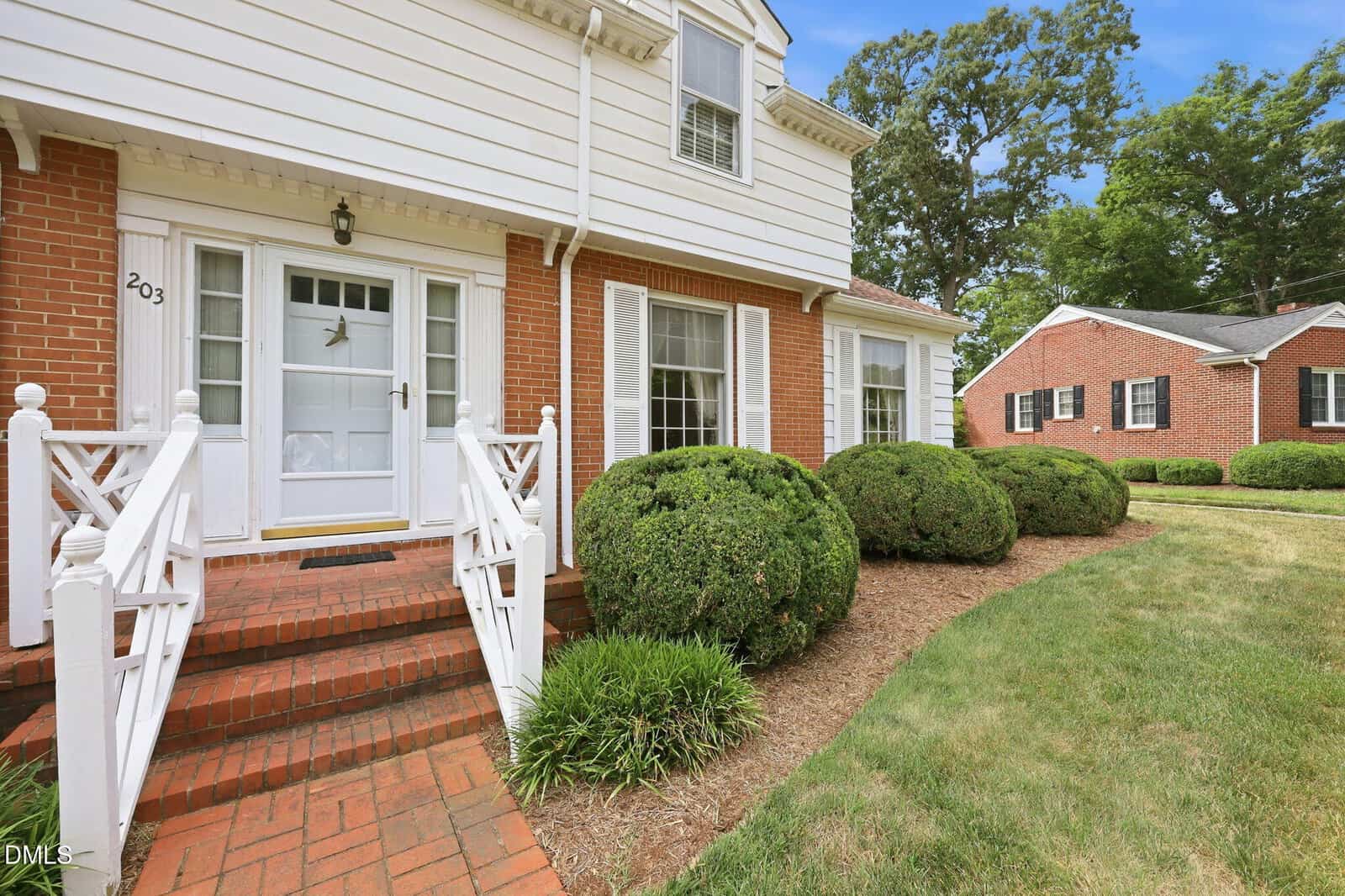 A brick house with white trim, a white front door, and shuttered windows, surrounded by neatly trimmed bushes and green grass, with another red brick building visible in the background.