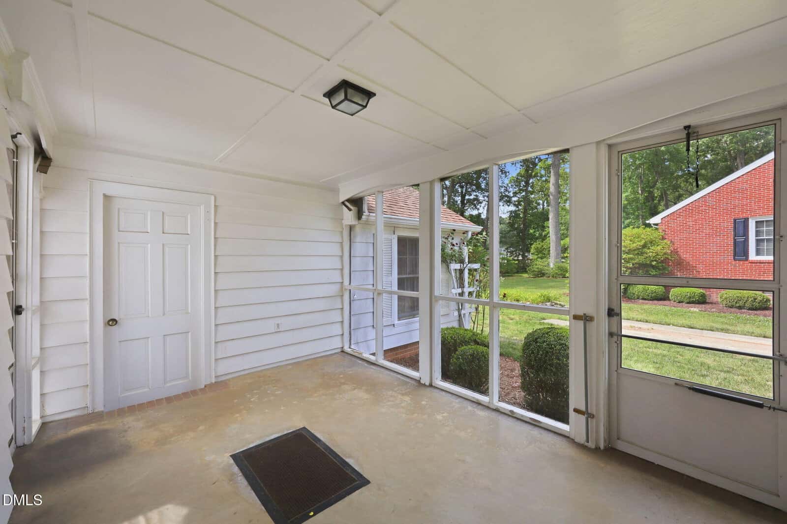 A screened-in porch with white paneled walls, a closed white door, and a concrete floor. Large screened windows offer a view of a green yard and a red brick house outside.