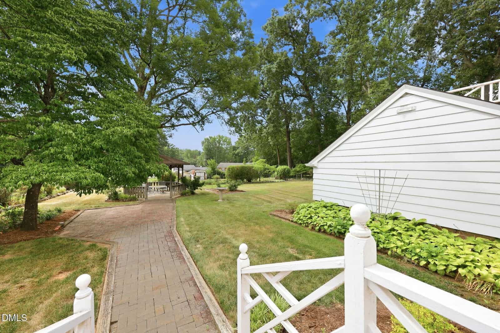 A paved walkway leads from a white-railed porch past a lawn and garden beds toward a gazebo and trees, with a white building on the right under a bright blue sky.