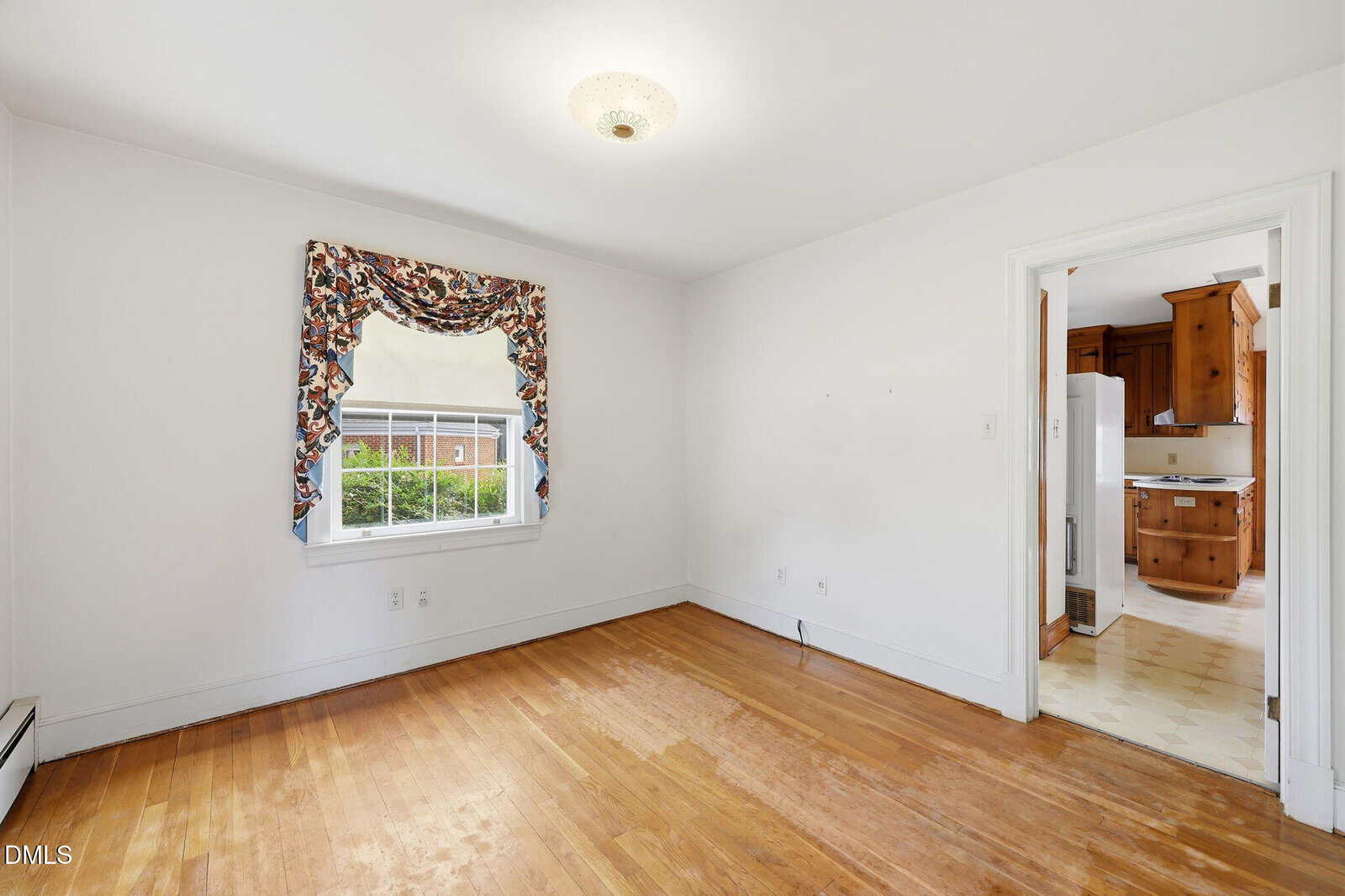 Empty room with hardwood floors, a window with a floral valance, white walls, and a ceiling light. An open doorway leads to a kitchen with wooden cabinets and visible white appliances.