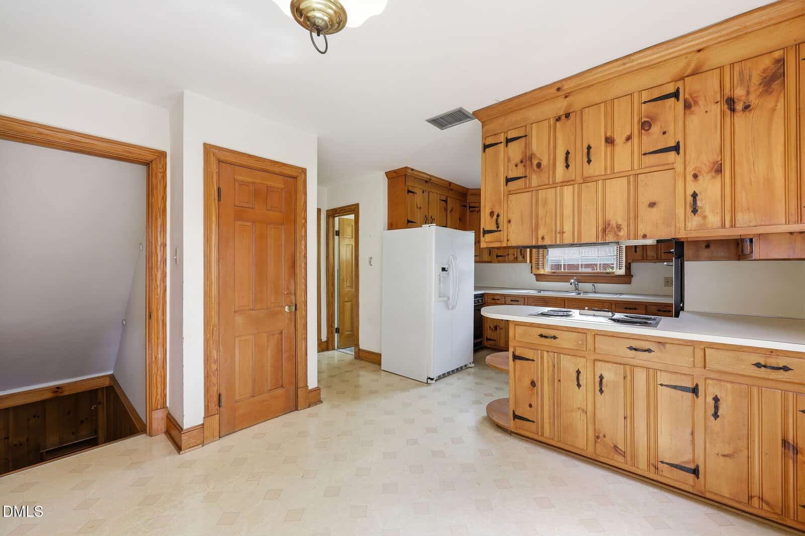 Kitchen with wood cabinets and doors, white appliances, beige patterned floor, and a small window above the sink. Staircase with wooden railing is visible to the left. The overall design has a rustic, vintage feel.
