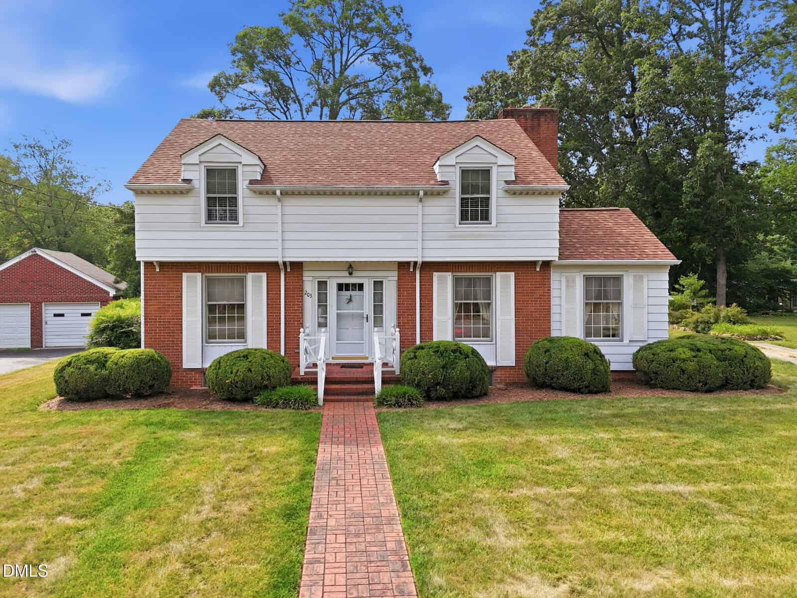 A two-story red brick and white siding house with dormer windows, a front porch, and a brick walkway, surrounded by green bushes and a neatly mowed lawn.