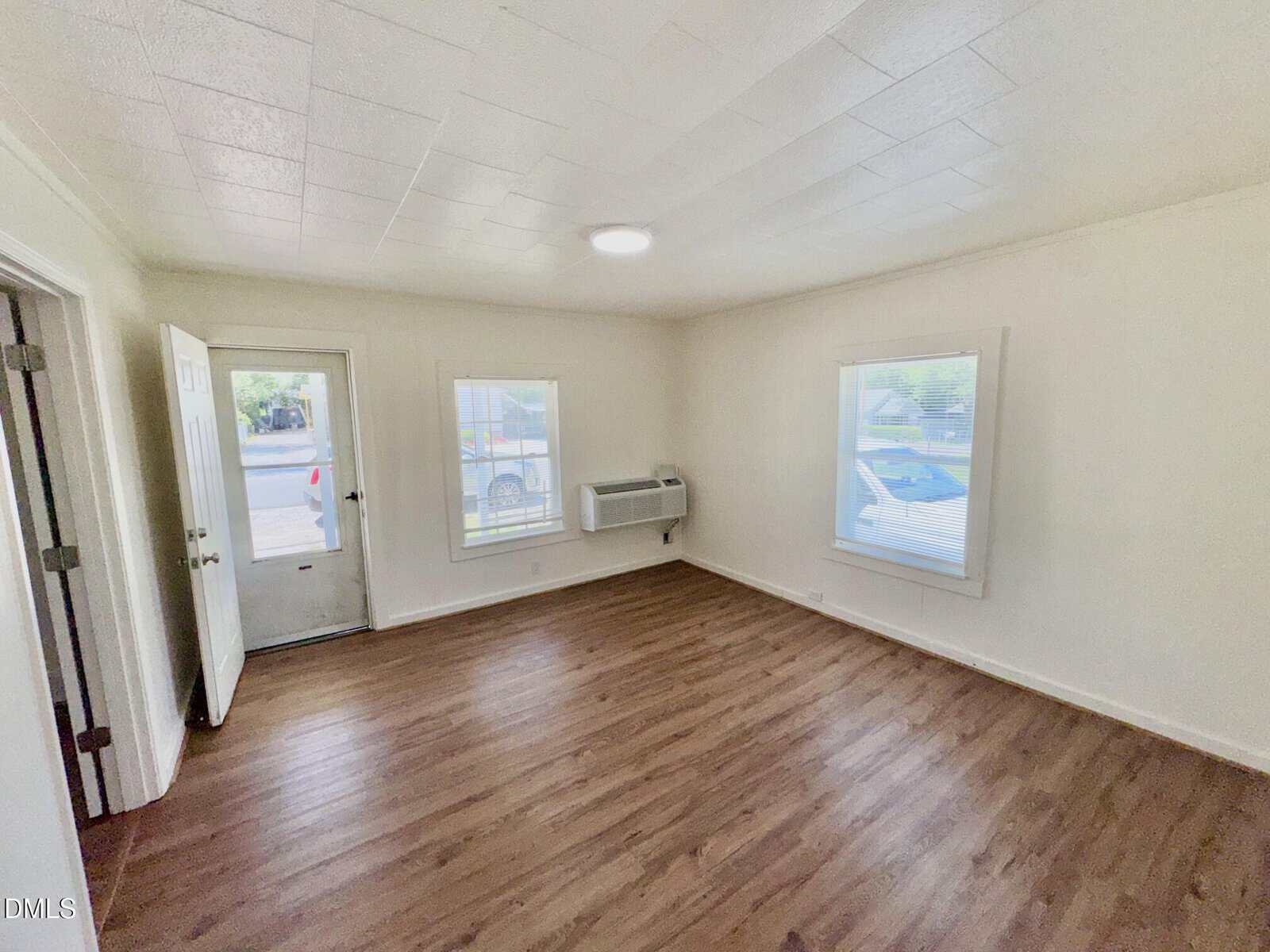 A bright, empty room with light-colored walls, wood flooring, two windows, a glass door, and a wall-mounted air conditioning unit. Natural light fills the space, creating an airy atmosphere.