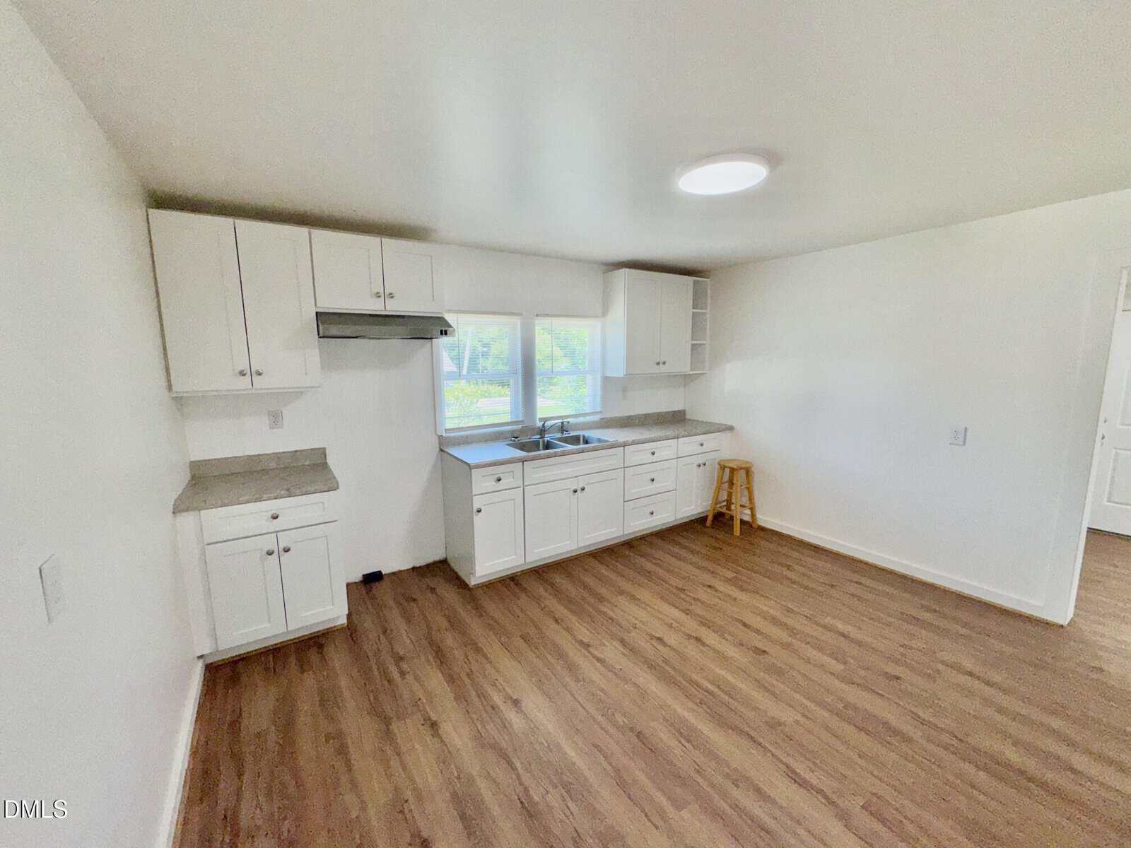 A bright, modern kitchen with white cabinets, gray countertops, a stainless steel sink under two windows, and wood-style flooring. A single wooden stool sits by the counter. The walls are painted white.