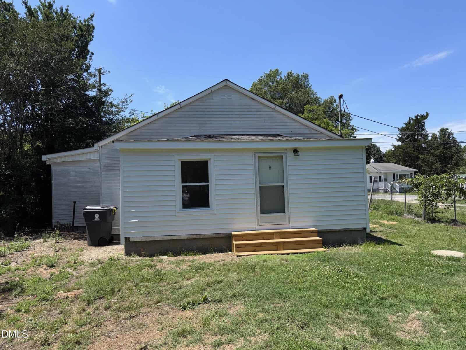 A small white single-story house with a simple porch and steps, surrounded by grass and a few trees. There is a trash bin on the left side and a neighboring house visible in the background. The sky is clear and blue.