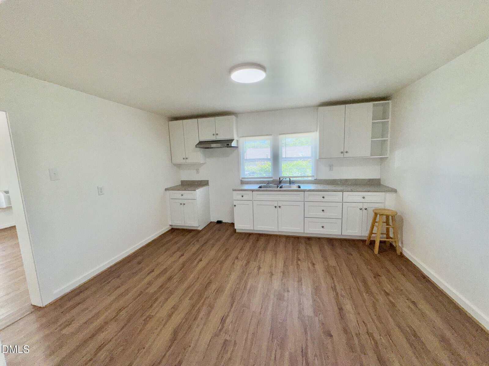 A small kitchen with white cabinets, a sink under two windows, wood flooring, and a single wooden stool near the counter. The room is brightly lit with a ceiling light.