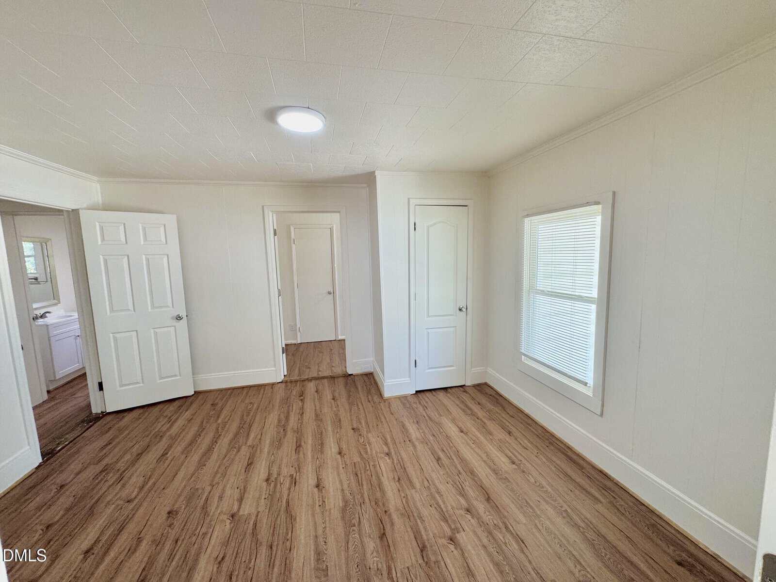 A bright, empty room with wood flooring, white walls, two closed white doors, and a large window with blinds letting in natural light.