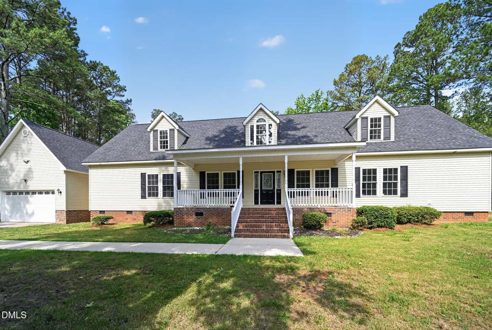 Located at 161 Sequoia Drive in Louisburg, this large two-story house features cream siding, black shutters, three dormer windows, a spacious front porch with white railings, brick steps, an attached garage, and a scenic lawn surrounded by trees.