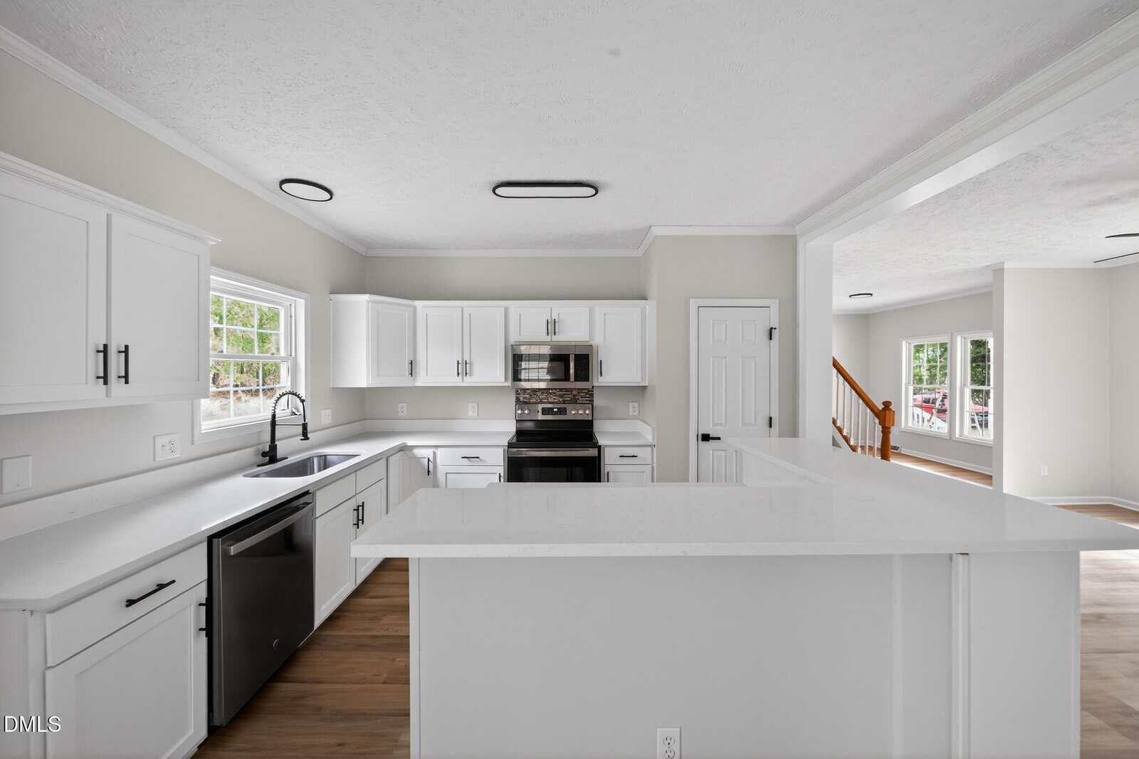 Modern white kitchen with shaker cabinets, stainless steel appliances, large sink, and bright windows. The open layout shows a view into the living area with wood floors and a staircase in the background.