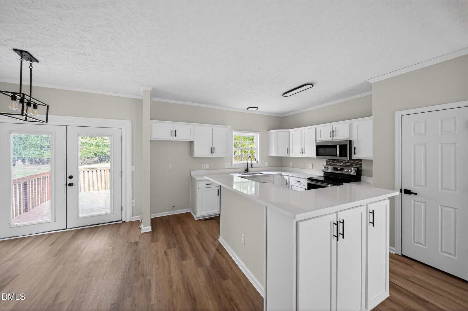 Bright modern kitchen with white cabinets, stainless steel appliances, and white countertops. Wooden floors, a window over the sink, and glass doors leading to a deck are also visible. The walls are light beige.