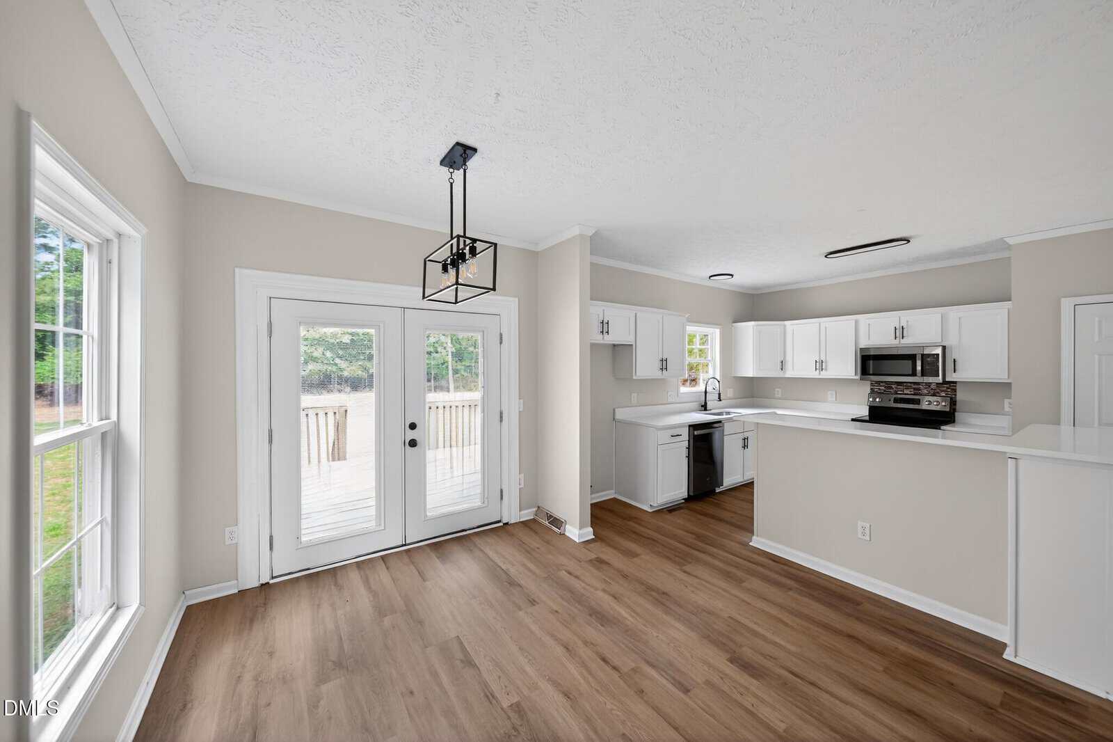 Bright kitchen and dining area with white cabinets, stainless steel appliances, light wood floors, large windows, and double glass doors leading to a backyard deck. The space features neutral walls and modern lighting.