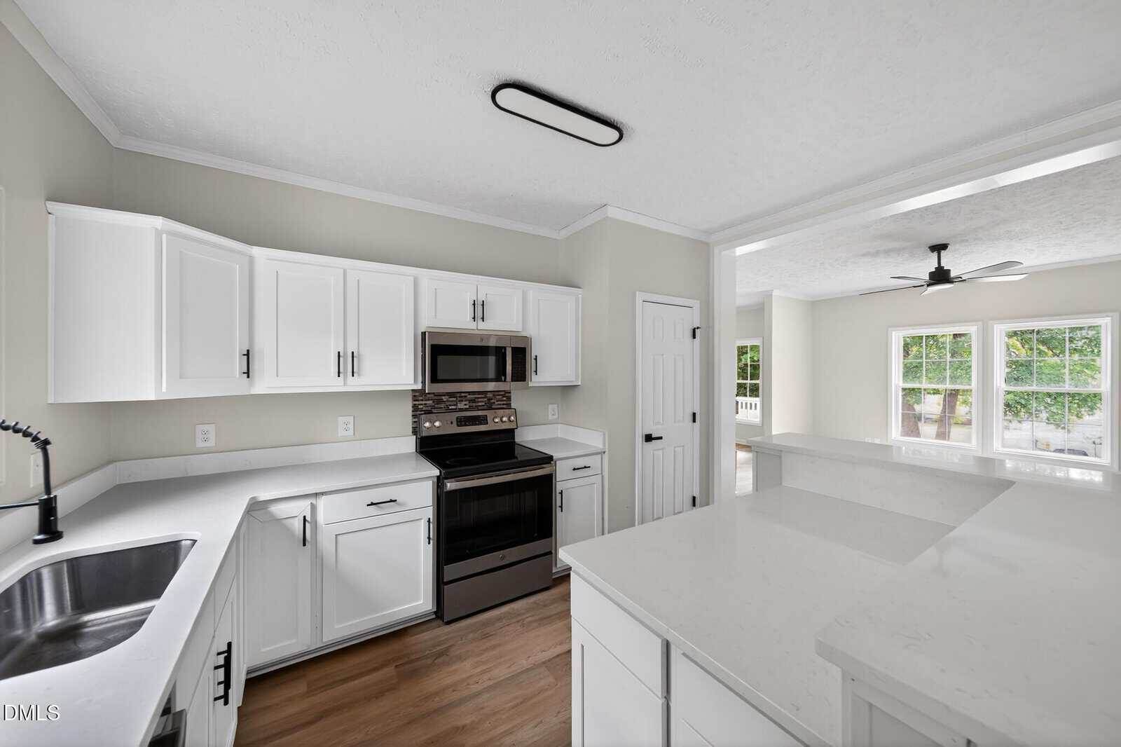 Modern kitchen with white cabinets, stainless steel appliances, a tile backsplash, and wood flooring. An open layout connects to a bright living area with large windows and a ceiling fan.