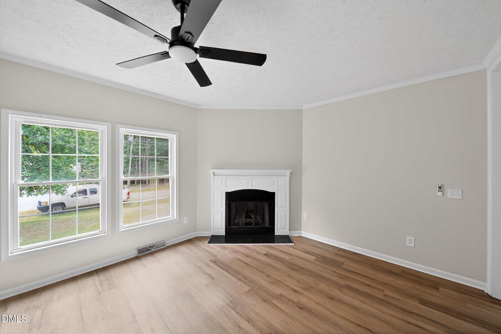 Bright, empty living room with light wood floors, white walls, black ceiling fan, two large windows, and a white fireplace centered on the far wall. Trees and a white vehicle are visible outside.
