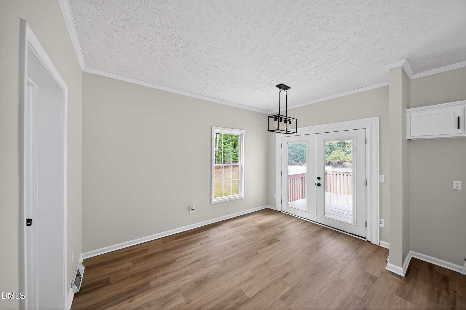 Bright, empty room with light wood flooring, neutral walls, a modern black chandelier, double glass doors leading to a deck, and a large window bringing in natural light.