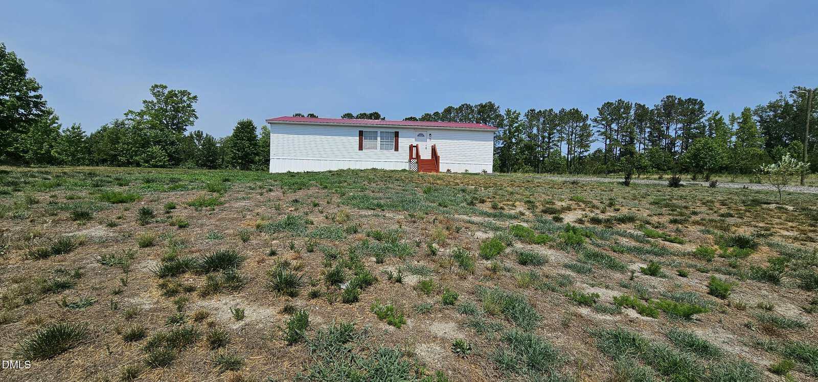 A small white house with a red roof and front steps sits alone on a large, dry, patchy grass field at 70 Waterfront Drive, Louisburg, surrounded by trees under a clear blue sky.