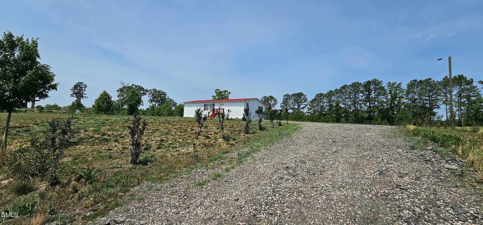 A gravel driveway leads up a slight hill to a white building with a red roof, surrounded by sparse grass, small trees, and a few larger trees in the background under a clear blue sky.