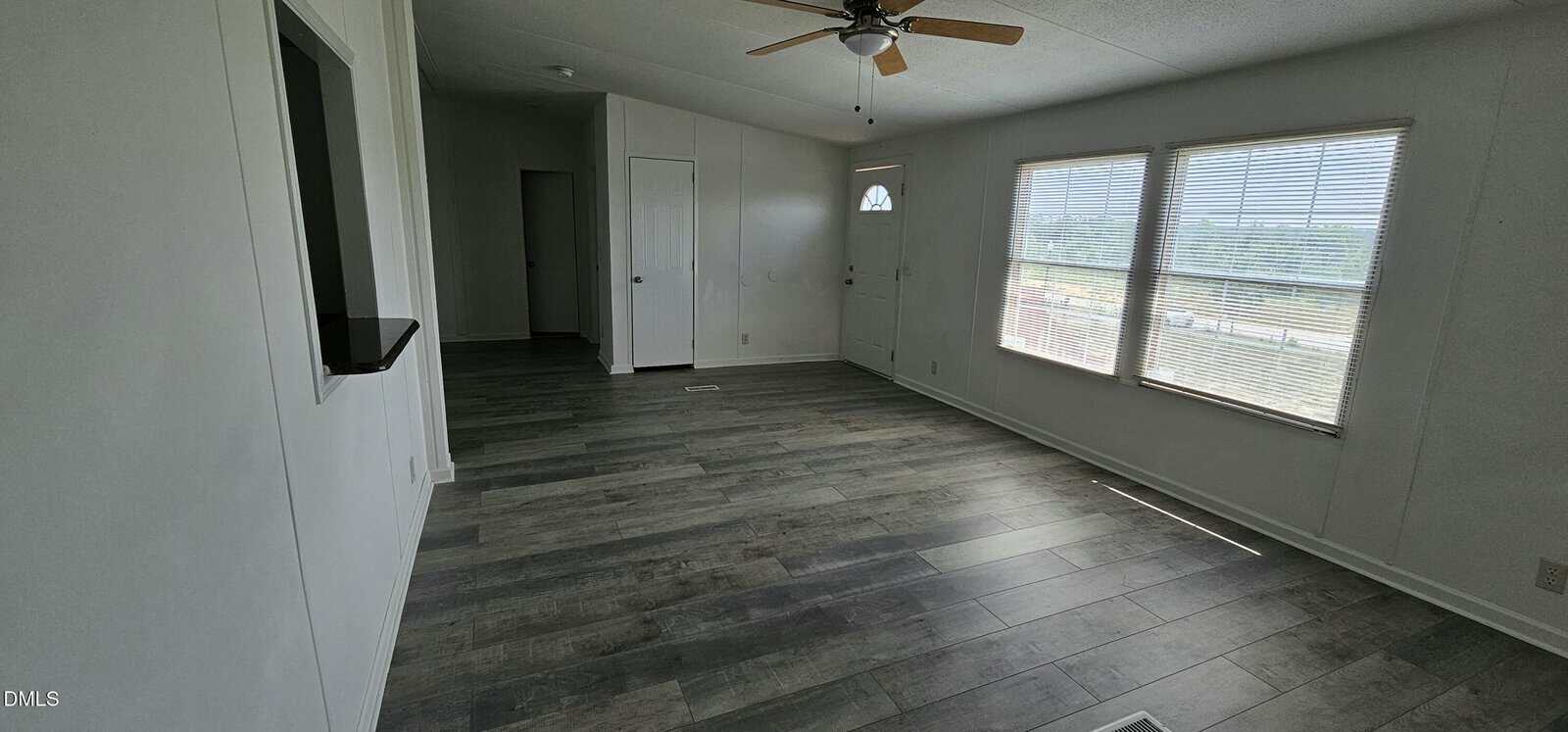 A bright, empty room with gray wood flooring, white walls, a ceiling fan, a door with a window, and three large windows letting in natural light. A small wall cutout opens to an adjacent area.