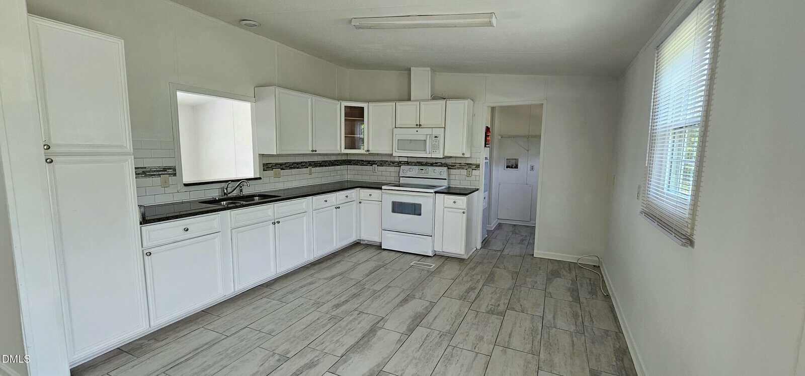Bright kitchen with white cabinets, appliances, and backsplash, gray tile floor, and large window on the right letting in natural light. A laundry area is visible through a doorway at the back.