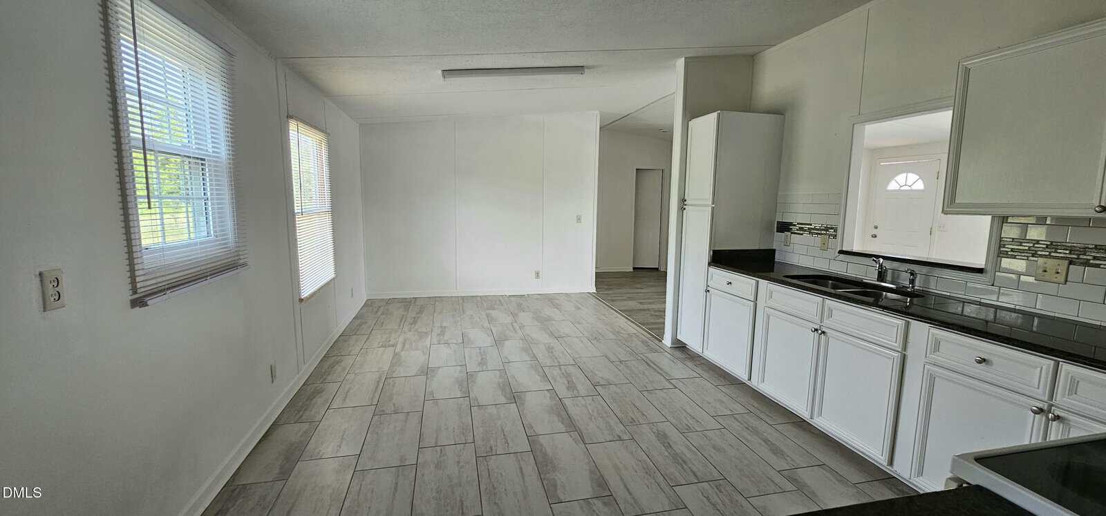 Bright kitchen with white cabinets and tiled floor, leading to an open dining area with large windows. A door and additional hallway are visible in the background.
