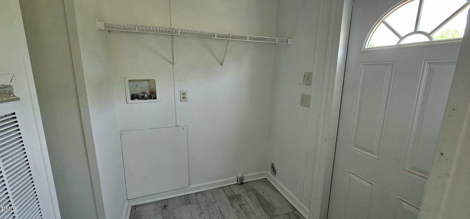 Small laundry area with white walls, a wire shelf, washer and dryer connections, electrical outlets, and a white door with a half-circle window on the right. The floor is covered with light gray tiles.