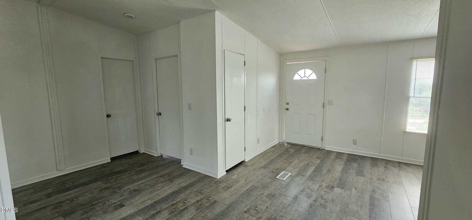 Bright entryway with white walls, three closed doors, a white front door with a half-moon window, and large window letting in natural light. Gray wood-style flooring throughout.