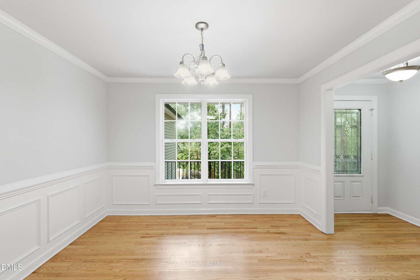A bright, empty dining room with white walls, wood floors, a large window showing greenery outside, and a door with glass panels. A five-light chandelier hangs from the ceiling.