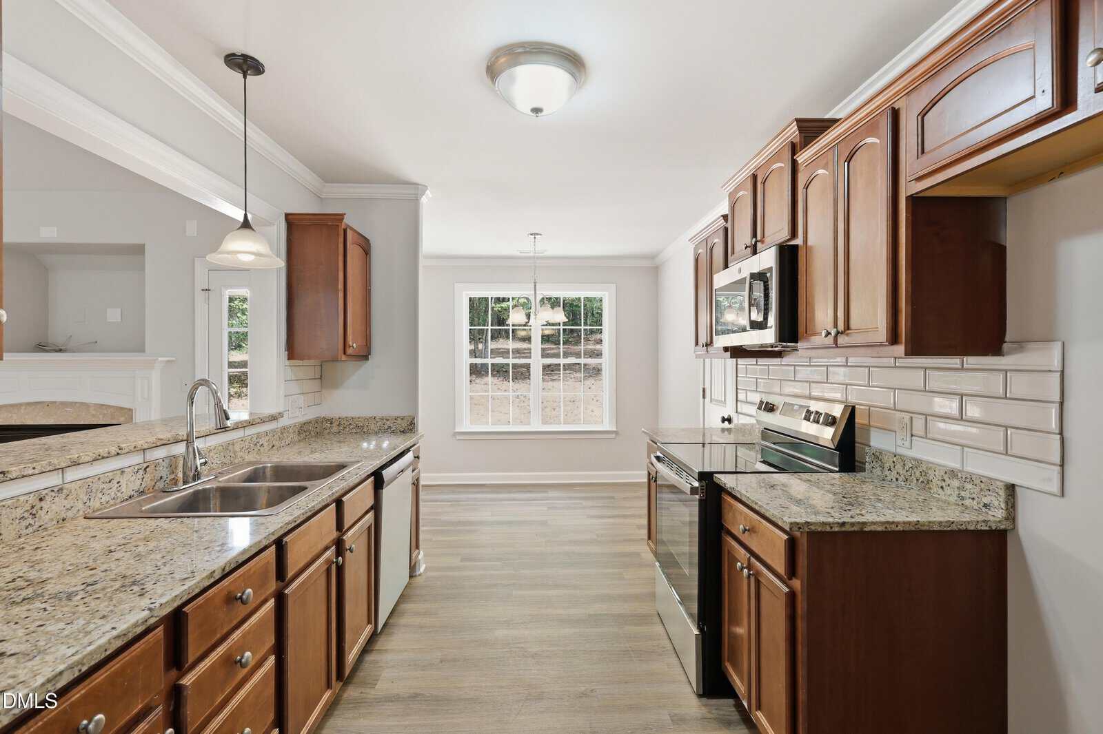 Modern kitchen with wooden cabinets, granite countertops, stainless steel appliances, tile backsplash, double sink, and a large window in the bright dining area with wood flooring. Walls are painted light gray.